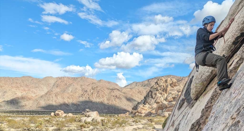 A person wearing safety gear is secured by ropes as they climb a rock wall. Behind them is a mountain range. 